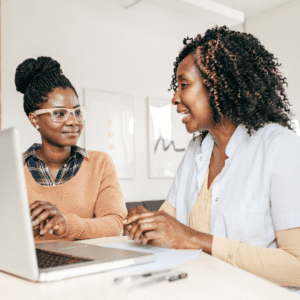 Two women engage in a collaborative discussion over a laptop in a bright office space, with one person appearing to provide guidance or consultation, fostering a professional yet warm atmosphere conducive to learning and support.