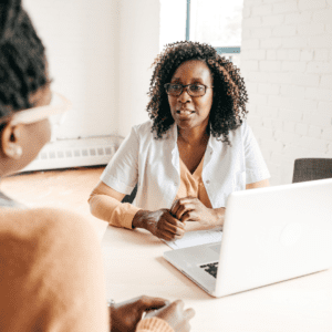 A consultant with curly hair, glasses, and a warm smile provides guidance during a one-on-one session, a laptop open for resources or note-taking, illustrating a personalized and professional approach to client care and education in a bright office setting.