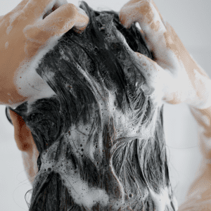 "Blog post visual from Alopecia & Beyond, dated March 13, 2024, with a close-up photo of a person massaging shampoo into their hair, paired with the headline 'Shampoo Ingredients That Cause Hair Loss'. The image invites readers to explore which shampoo components might be detrimental to hair health