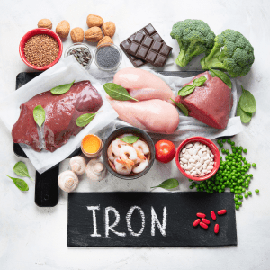 Promotional graphic for a blog post titled 'The Benefits of Iron For Hair Growth' from Alopecia & Beyond, dated March 13, 2024. Features an assortment of iron-rich foods and a chalkboard labeled 'IRON', with a call-to-action prompting to 'Read More' about iron's impact on hair health.